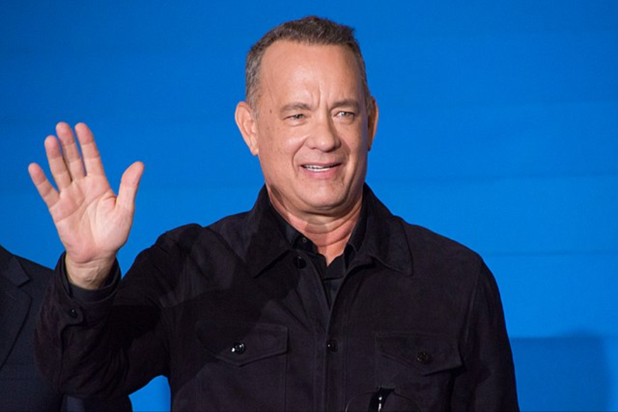 Man waves with a friendly smile while wearing a dark jacket in front of a blue background.