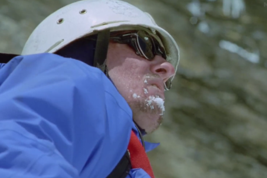 A close-up of a climber with frost on his face wearing glacier goggles and a helmet, looking up while surrounded by snow and ice.