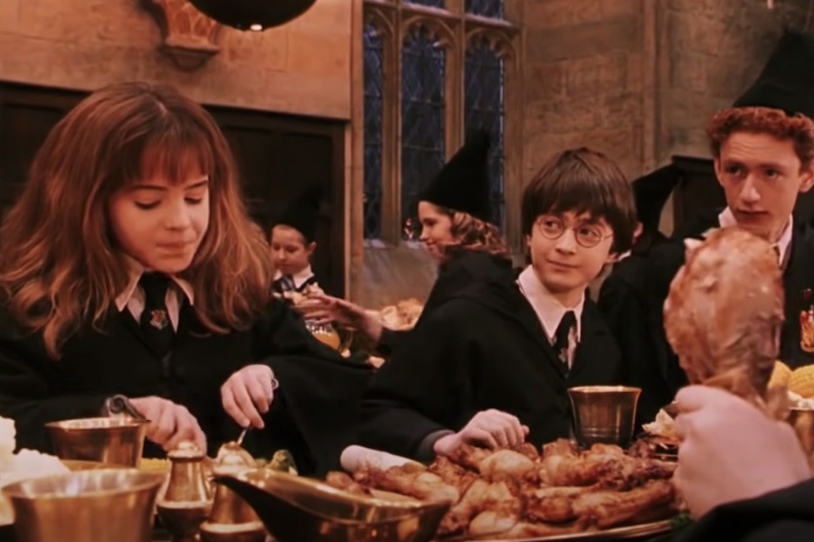 Three young students in wizard robes eat a feast in a candlelit dining hall, with a girl focused on her plate and a boy holding a large turkey leg.