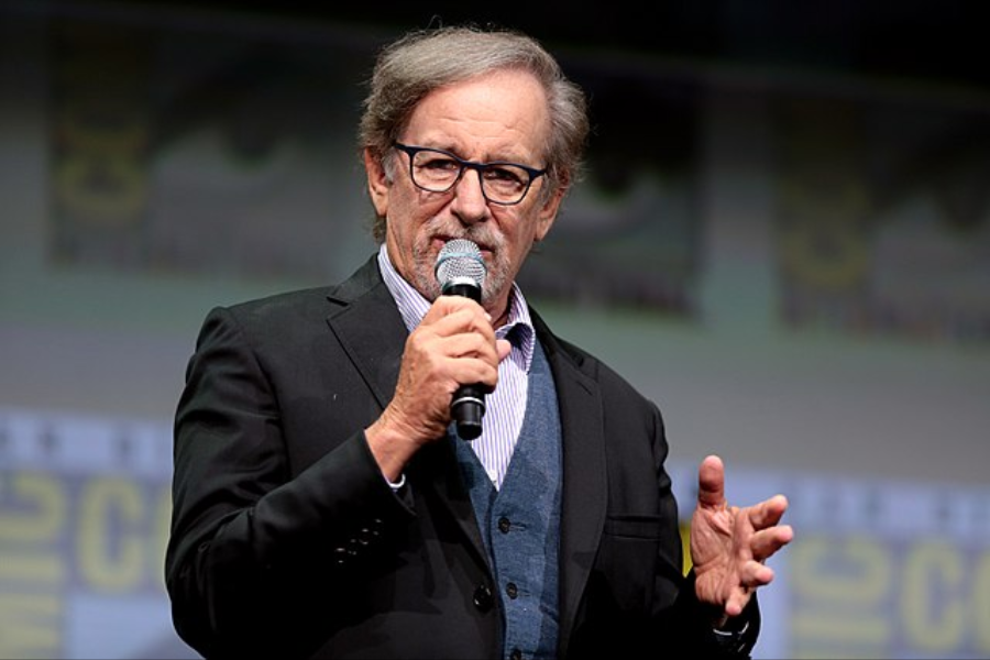 Man in a black suit and glasses speaks into a microphone on stage at a large conference or convention.