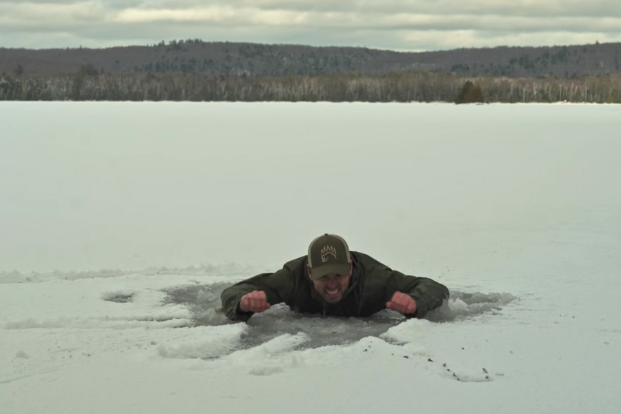 Man emerging through a hole in a frozen lake, clenching his fists and grimacing in the middle of a vast snow-covered landscape.