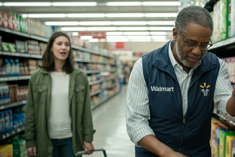 Walmart employee wearing glasses and a blue vest works in an aisle while a woman in a green jacket speaks behind him.