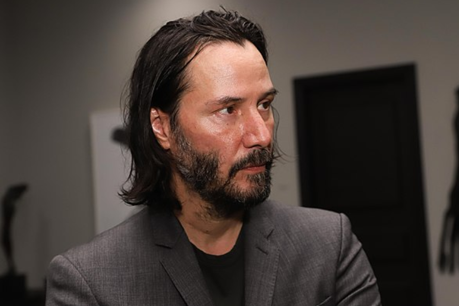 Bearded man with shoulder-length hair listens intently while wearing a grey blazer in an indoor setting.