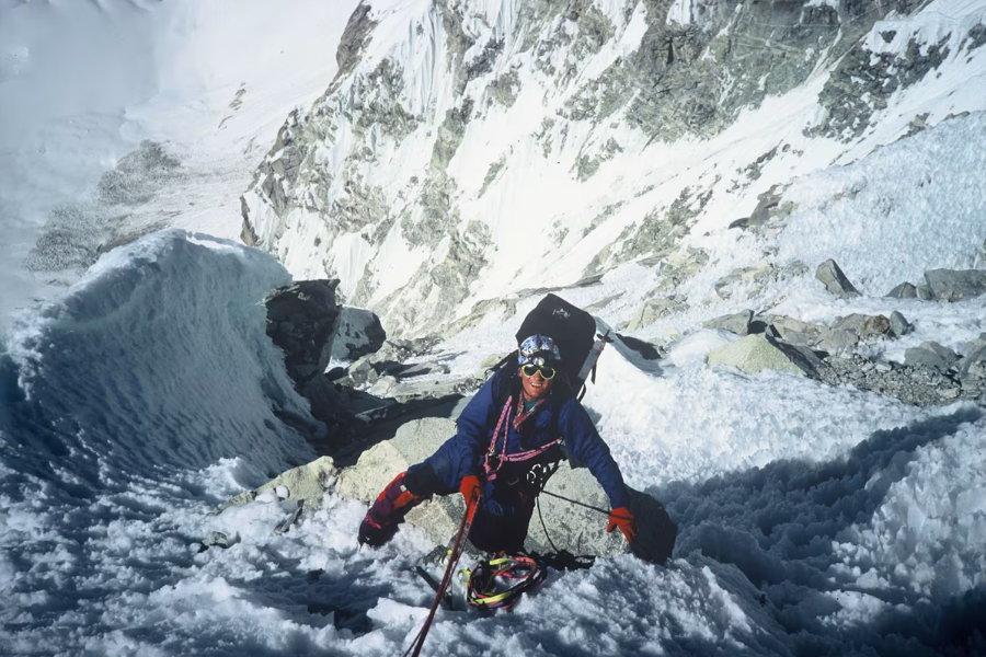 A mountaineer in bright gear climbs a steep, icy ridge, secured by rope, with jagged, snow-covered peaks stretching below and behind.