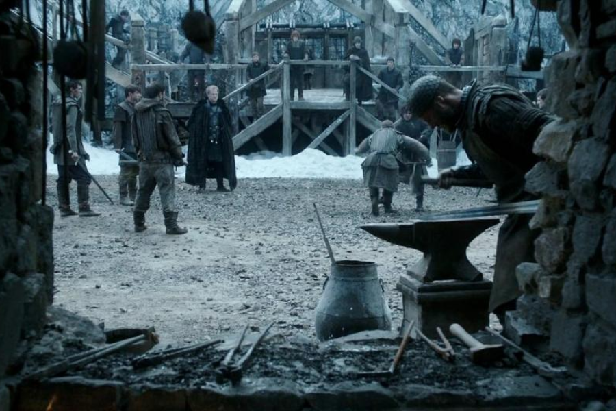 A blacksmith works at an anvil in the foreground while people gather in a snowy training courtyard in a medieval setting.