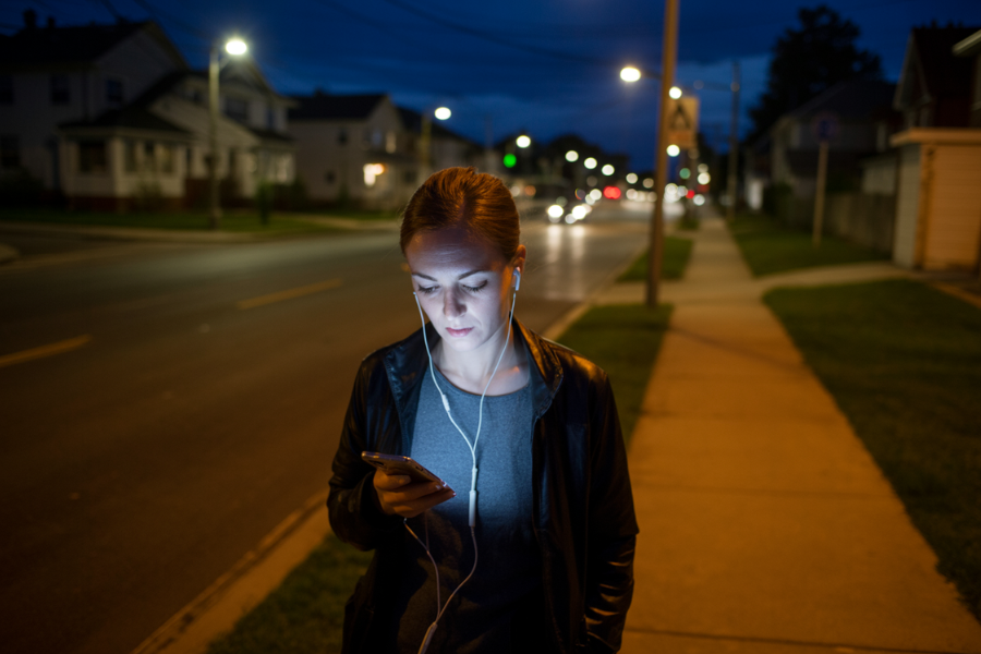 Woman walking on a quiet residential sidewalk at night, illuminated by the glow of her smartphone screen while wearing earphones.