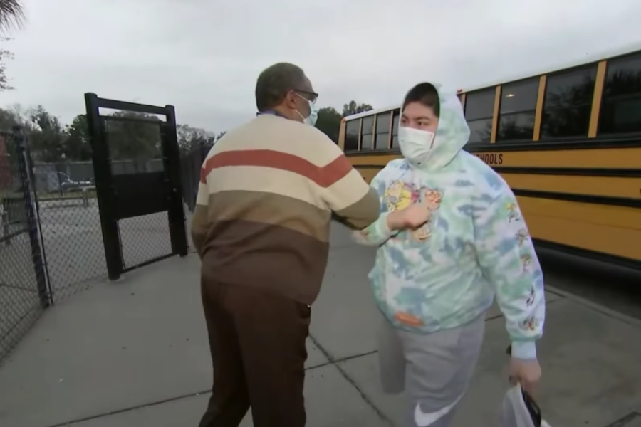 Man in a striped sweater stops a masked student wearing a colorful hoodie near a yellow school bus.