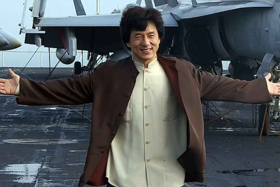 Man with arms outstretched smiles broadly while standing on an aircraft carrier deck in front of fighter jets.