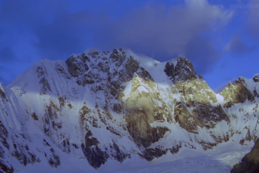 A sunlit snow-covered mountain peak rises sharply under a twilight sky, with dramatic shadows cast across its icy face.