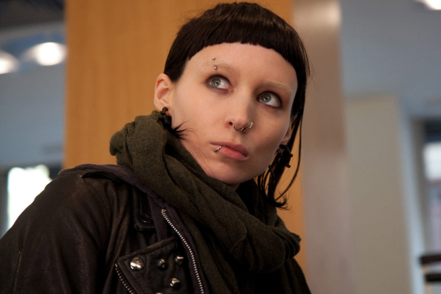 A young woman with dark makeup, piercings, and a blunt haircut looks upward in a modern indoor setting.