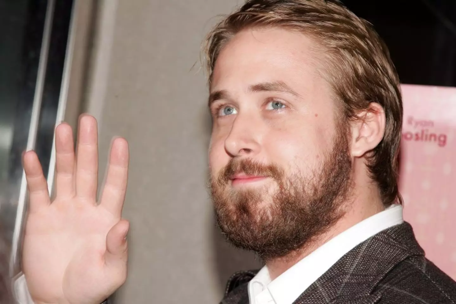 A bearded man with light brown hair and a suit raises his hand in a subtle wave, standing in front of a pink backdrop.