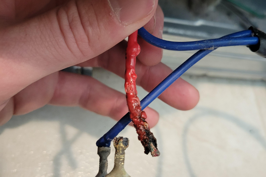 Close-up of a burnt red electrical wire with exposed damage held next to intact blue wires, showing signs of overheating or short-circuiting.