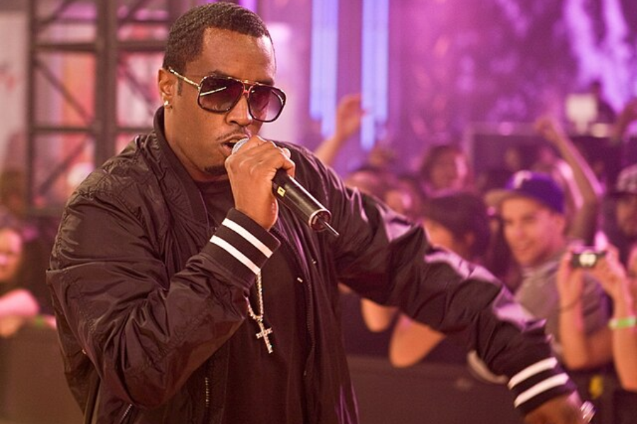 Performer in sunglasses and a black jacket raps into a microphone on stage with a cheering crowd behind him.