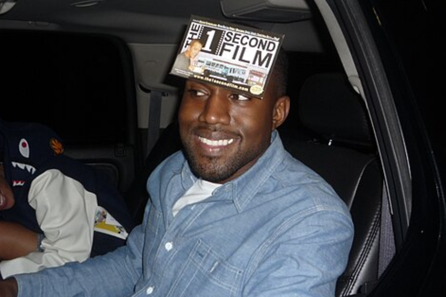 Man sitting in a car smiling with a promotional flyer stuck to his forehead that reads “The 1 Second Film.”