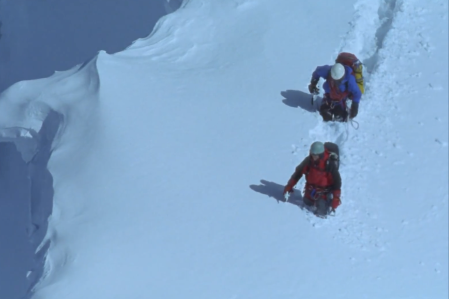 Two climbers trek through deep snow, viewed from above, with a sharp cornice on the left and a clear trail of their ascent behind them.