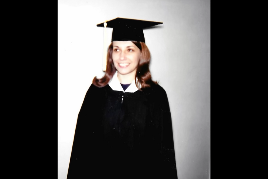 Young woman in a black graduation cap and gown smiling toward the side. She stands against a plain light background.