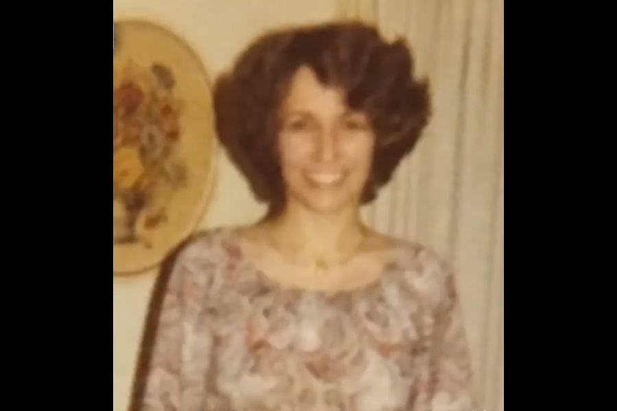 Vintage indoor photo of a woman with curly hair wearing a patterned blouse. She smiles while standing in front of light curtains with a floral wall plate nearby.