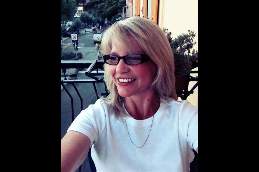 A woman with blonde hair and glasses smiles while taking a selfie outdoors on a balcony overlooking a street.