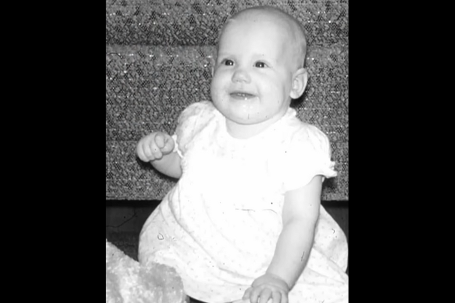 Smiling baby in a light dress sitting on the floor in front of patterned carpeted stairs.