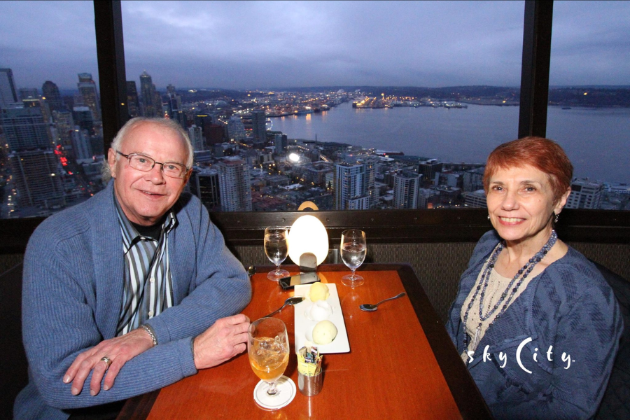 A couple sits at a restaurant table by a large window overlooking a city and harbor at dusk.