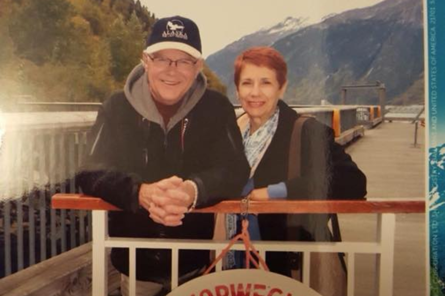 A man in a cap and hoodie and a woman in a red coat smile while standing on a dock with snowy mountains in the background.