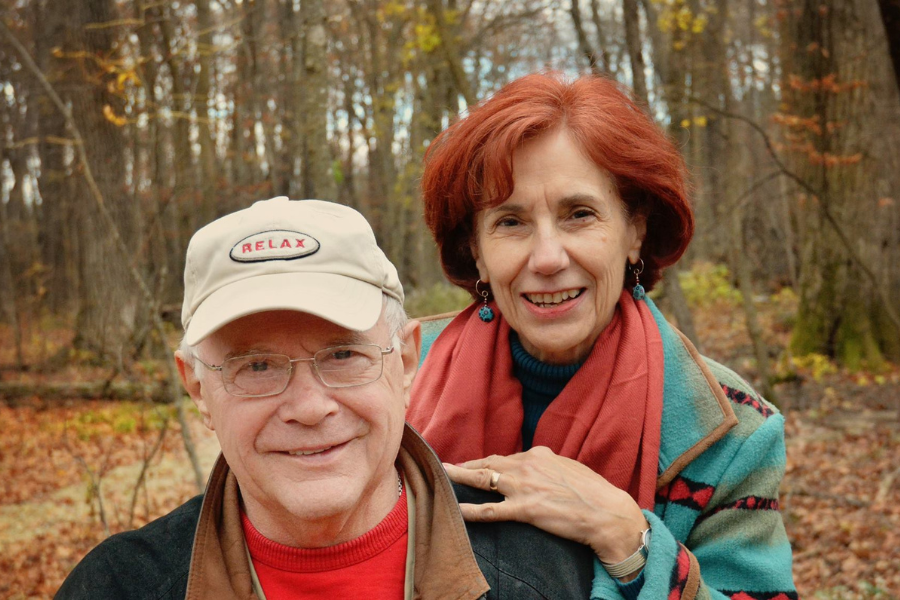 A smiling couple poses outdoors in the woods during autumn, wearing cozy jackets and scarves.