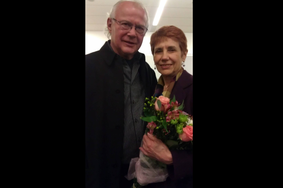 An older couple poses indoors, the woman holding a bouquet of flowers while the man stands beside her with his arm around her.