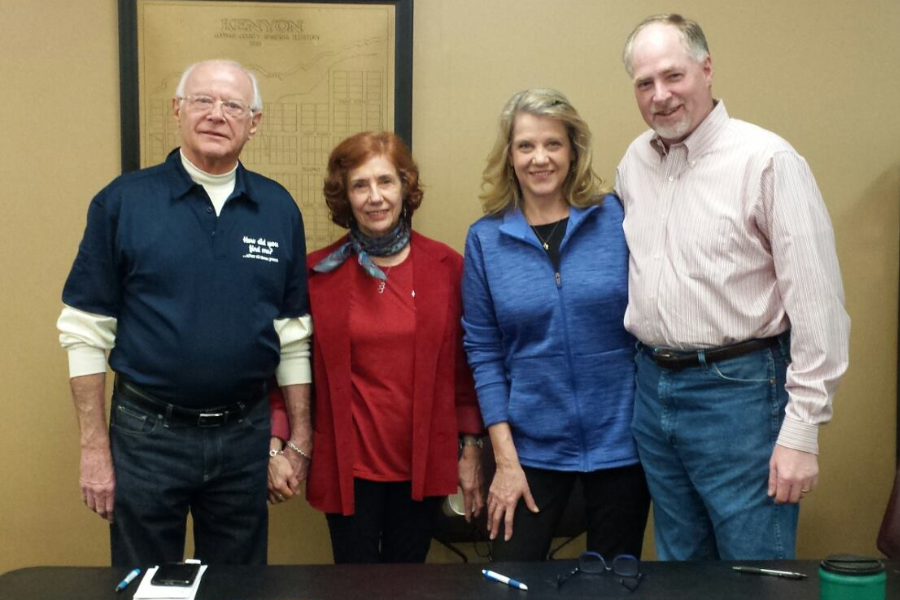 Four adults stand in a row indoors, smiling at the camera, with a table and papers in front of them.