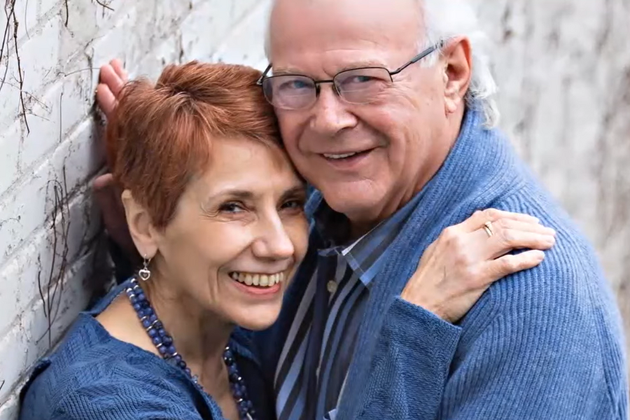 An older couple smiles warmly while embracing against a brick wall, both dressed in blue sweaters.