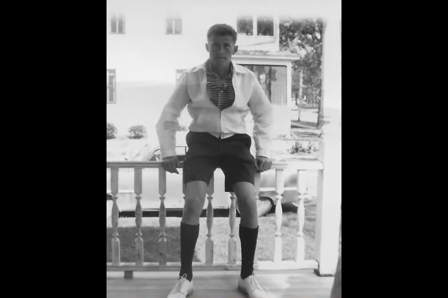 Black and white photo of a teenage boy sitting on a porch railing wearing a light jacket a striped shirt shorts knee high socks and white sneakers. A house and trees are visible across the street.
