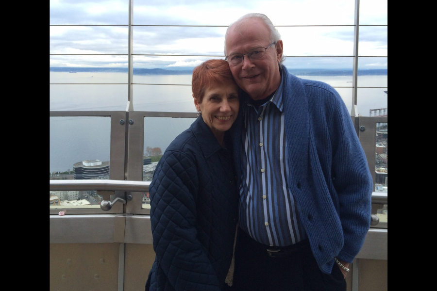 A couple embraces on a tall outdoor observation deck overlooking water and city buildings.