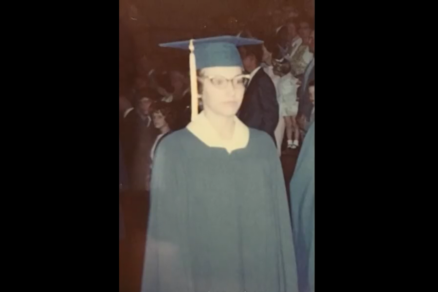 Faded color snapshot of a graduate in a teal cap and gown walking in a crowded auditorium. The person wears glasses and looks forward while people stand in the background.