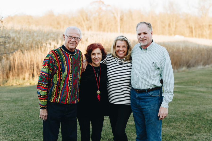 Four adults stand close together outdoors in a field, smiling, with autumn trees in the background.