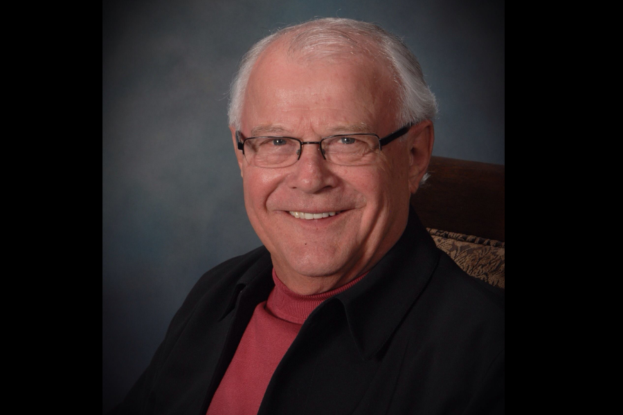 Studio headshot of an older man with white hair glasses and a warm smile wearing a maroon turtleneck and dark jacket. The background is a soft blue gray gradient.