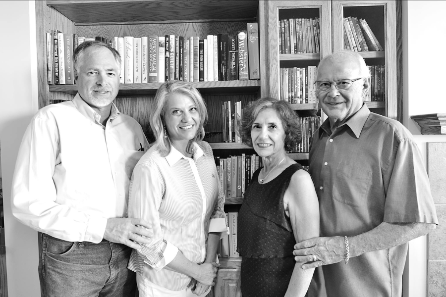 Black and white photo of two couples standing in front of a bookshelf, smiling for the camera.