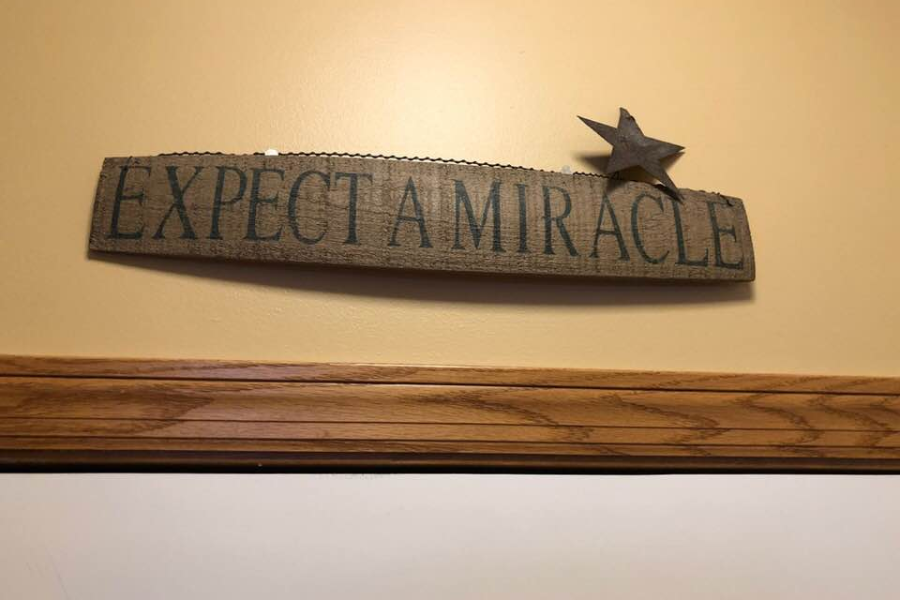 Wooden wall sign with a small metal star reads EXPECT A MIRACLE. The sign hangs above wood trim on a tan wall.