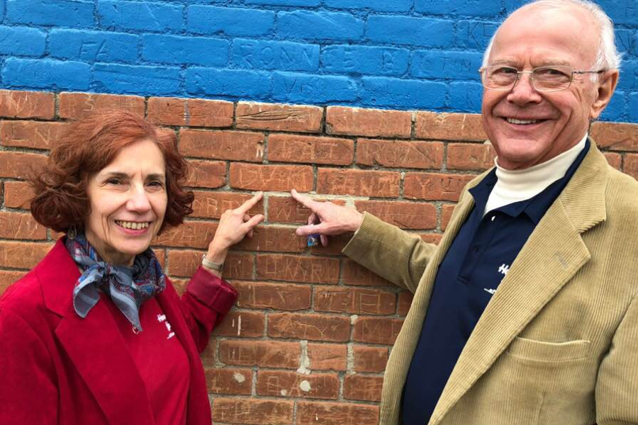 A smiling couple points to their carved initials on a brick wall painted partly blue.