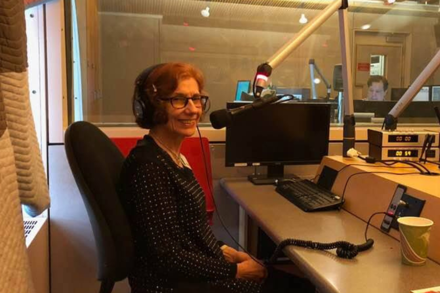 A woman with red hair and glasses smiles while sitting in a radio studio, wearing headphones with recording equipment and a computer in front of her.
