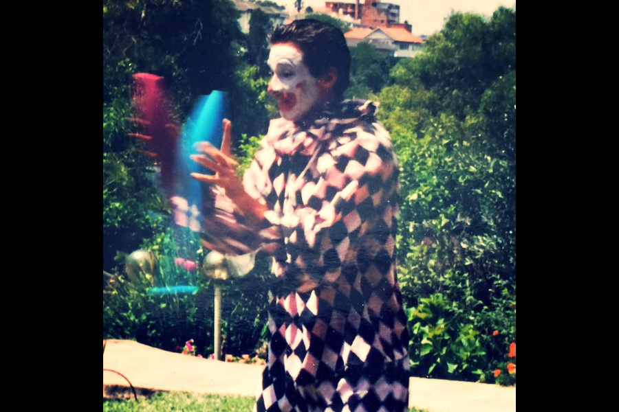 Clown in checkered costume and white face paint juggles colorful pins outdoors amidst lush trees and distant buildings, evoking a joyful scene.