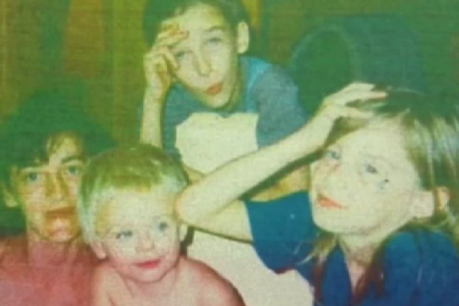 Four children sit closely together indoors, smiling and making playful gestures with their hands.