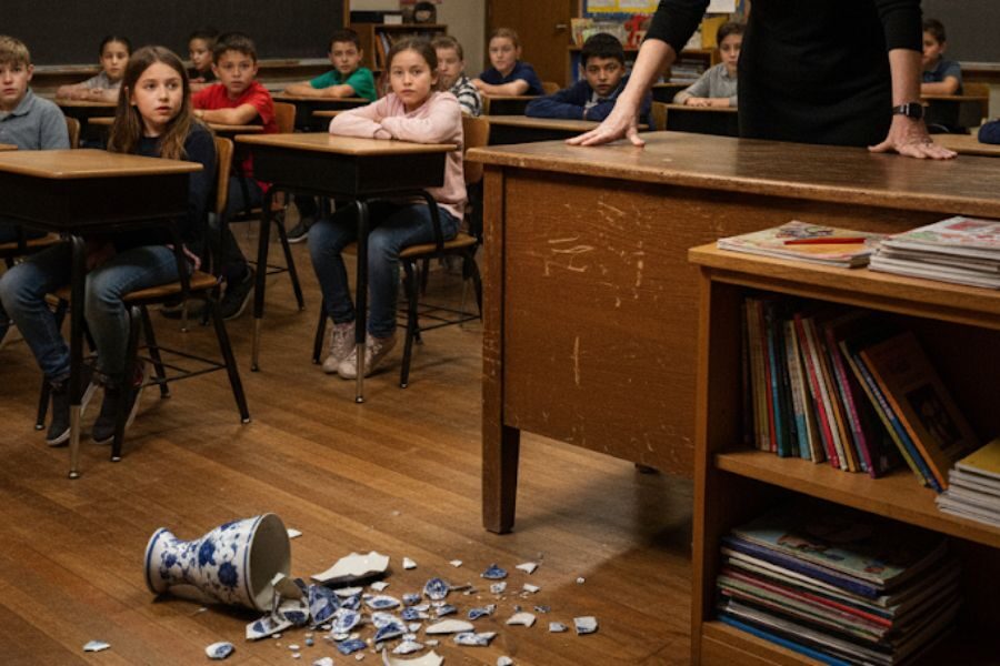 A broken blue and white porcelain vase lies shattered on the floor of a classroom as young students look toward the teacher, who stands at the front desk with a stern posture.