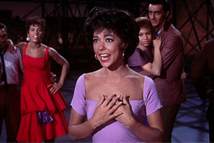 A woman in a lavender dress stands center stage, singing with her hands on her chest while others in vibrant outfits watch her in the background. The scene is theatrical and expressive with dramatic lighting.