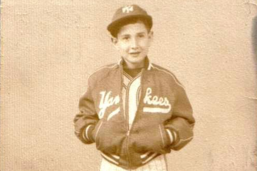 Alt text: A sepia-toned photo of a young boy wearing a Yankees jacket and cap, standing with his hands in his pockets and looking confidently at the camera.