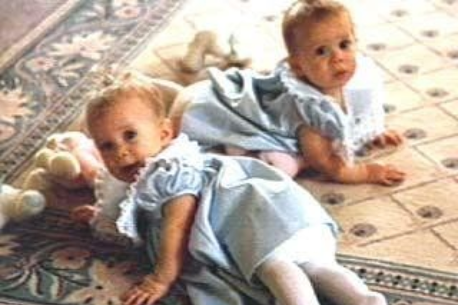 Twin babies lie on a patterned carpet in similar blue dresses with white lace collars, surrounded by plush toys, looking curiously at the camera.