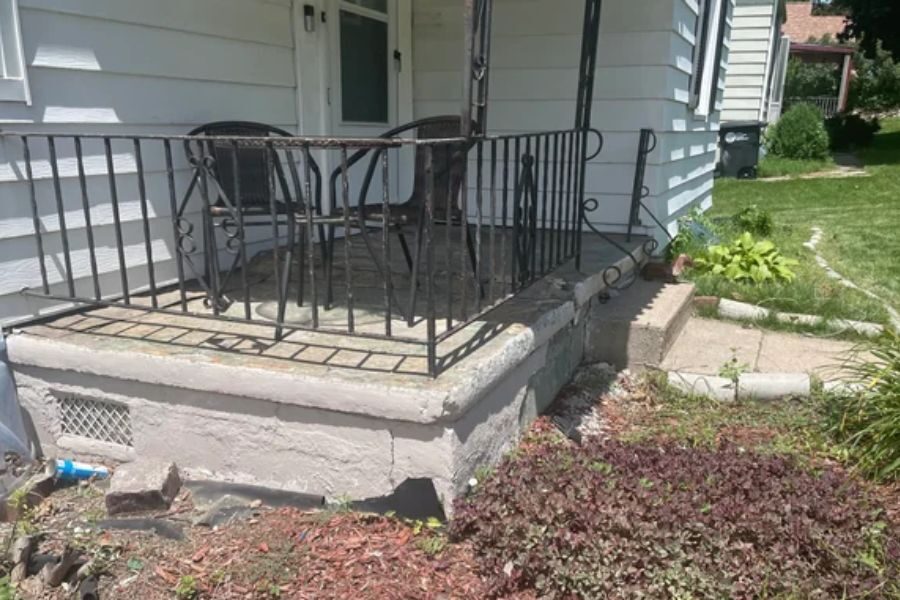 Small concrete porch with a metal railing and two chairs, attached to a white-sided house with a garden.