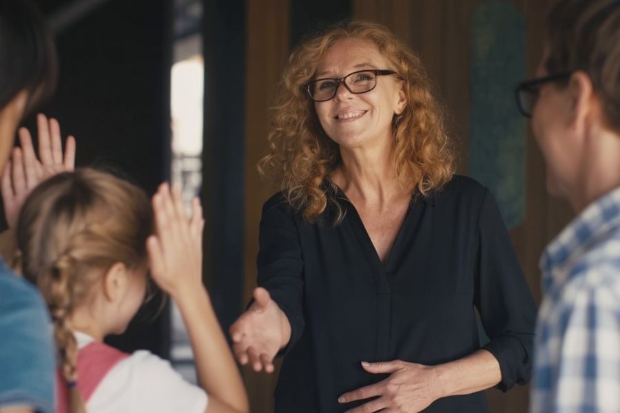 A smiling woman with curly hair and glasses extends her hand for a handshake while children raise their hands to greet her.