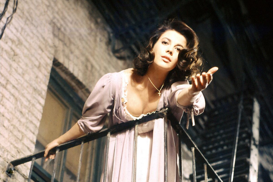 A woman in a flowing lavender nightgown stands on a fire escape balcony at night, reaching her hand out with emotion toward someone unseen below.