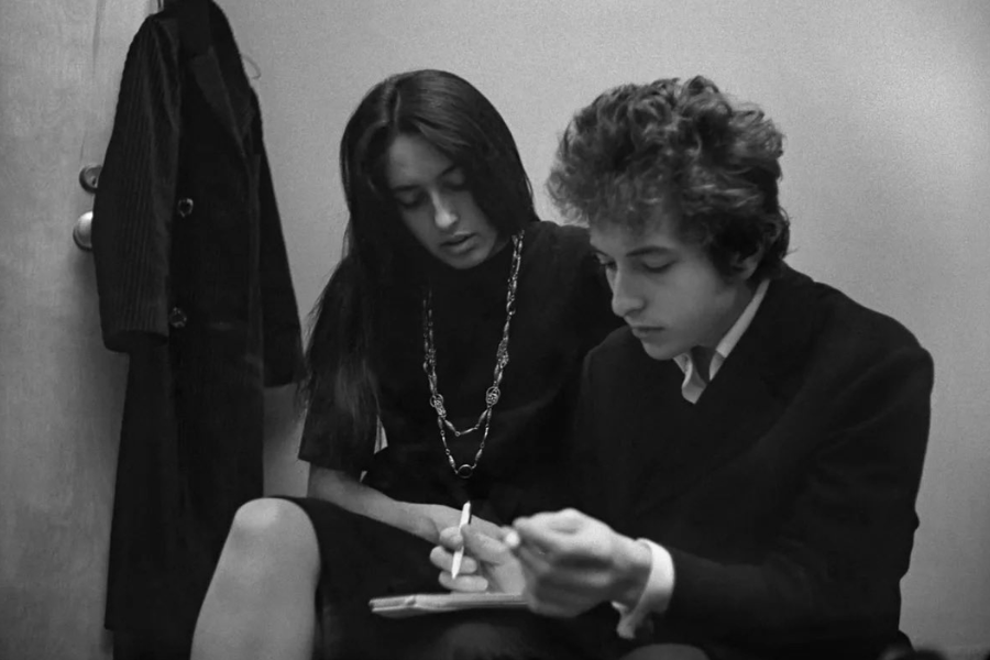 Intimate backstage photo of Joan Baez and Bob Dylan seated, with Baez watching Dylan write in a notebook.
