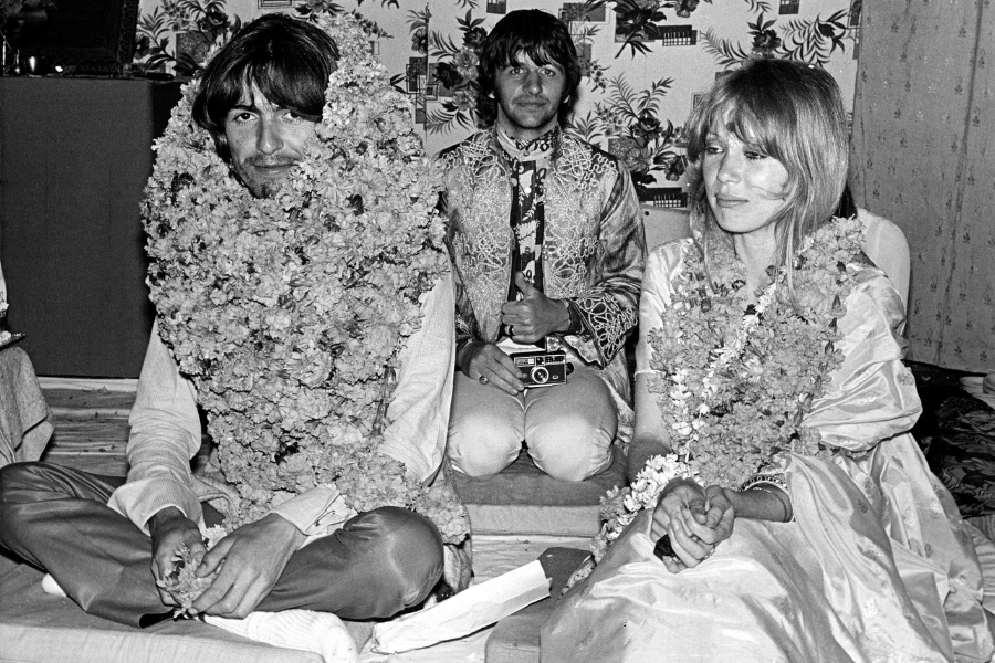 George Harrison and Pattie Boyd seated in traditional Indian attire and covered in flower garlands, with Ringo Starr sitting behind them holding a camera.