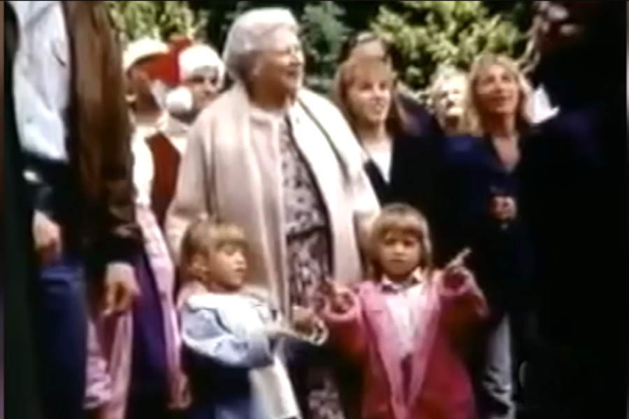 Two young girls stand among a group of smiling adults during a holiday gathering. Santa Claus characters and festive decorations are visible in the background.